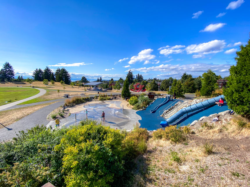 Sunny day at Jefferson Park in Seattle, showcasing kids playing on colorful slides and splashing in the spray pad, with open grassy fields and blue skies—one of the best Seattle parks to visit with kids for outdoor fun.