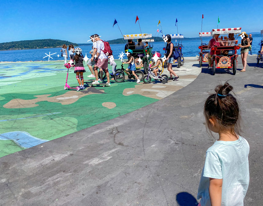 Point Ruston Waterfront is a family-friendly destination in Tacoma featuring wide, car-free paved walkways ideal for kids biking or riding scooters. In this photo, children ride alongside their family while enjoying the scenic Puget Sound views. Families can bring their own bikes and scooters or rent them from nearby vendors. With its safe pathways and fun atmosphere, Point Ruston is a must-visit spot for any Seattle-area family vacation itinerary