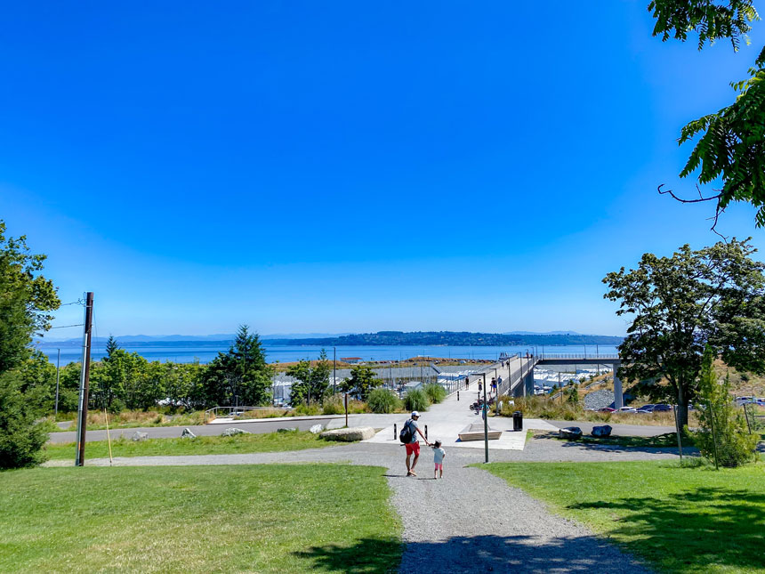 Part of our family-friendly guide to the Stairs and Slides in Tacoma—if you park at Point Defiance Park, you can enjoy a scenic and easy walk to the playground area. In this photo, a toddler and dad walk hand-in-hand along the paved path toward the hillside slides, with the added bonus of being near Point Ruston’s waterfront shops, dining, and splash pad. The stroll through the park is stroller-friendly and perfect for young kids, making it a great way to combine nature, play, and family bonding all in one outing.