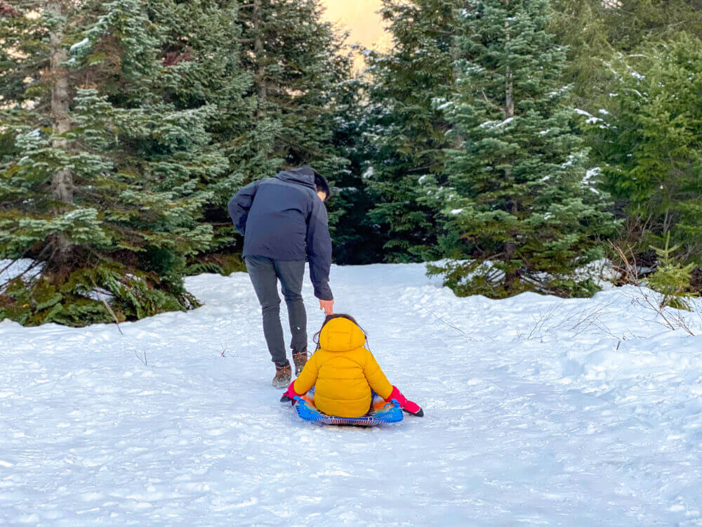Dad pulling his bundled-up child on a sled along a snow-covered trail at Gold Creek Pond during winter in Snoqualmie Pass. Surrounded by snowy trees and stunning mountain views, this scenic and family-friendly destination offers flat, easy trails perfect for winter walks, snowshoeing, and sledding with young kids—making it a peaceful escape for families near Seattle.
