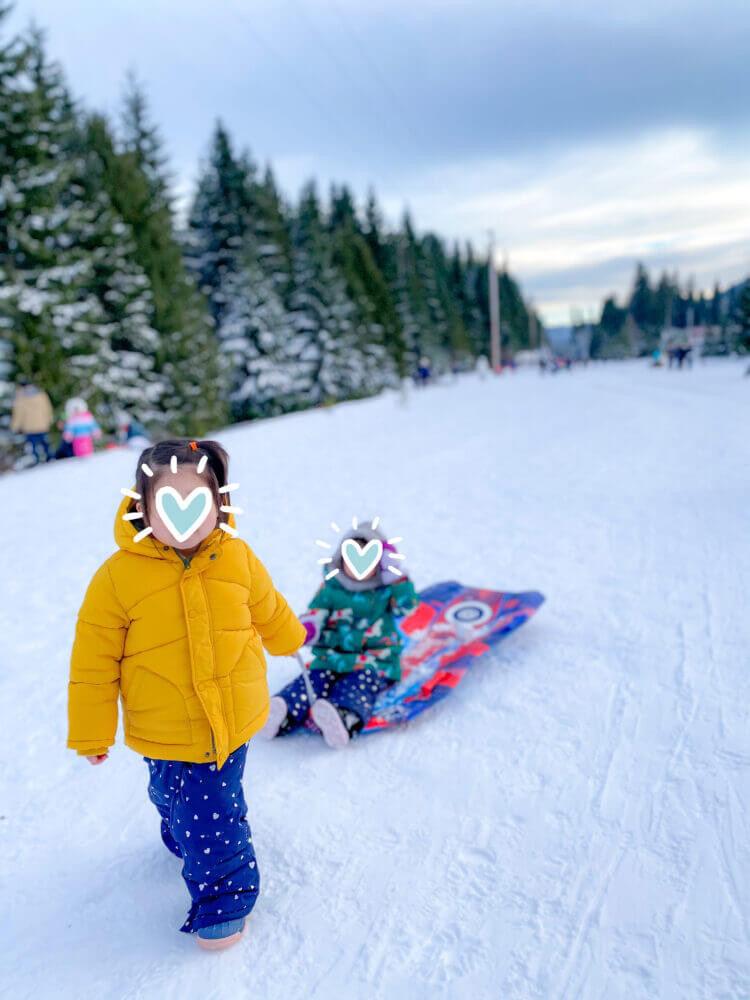 Young toddler girl pulling her friend on a sled through the snow at Hyak Sno-Park, a popular family-friendly winter destination near Seattle known for its scenic sledding hills and snowy fun.