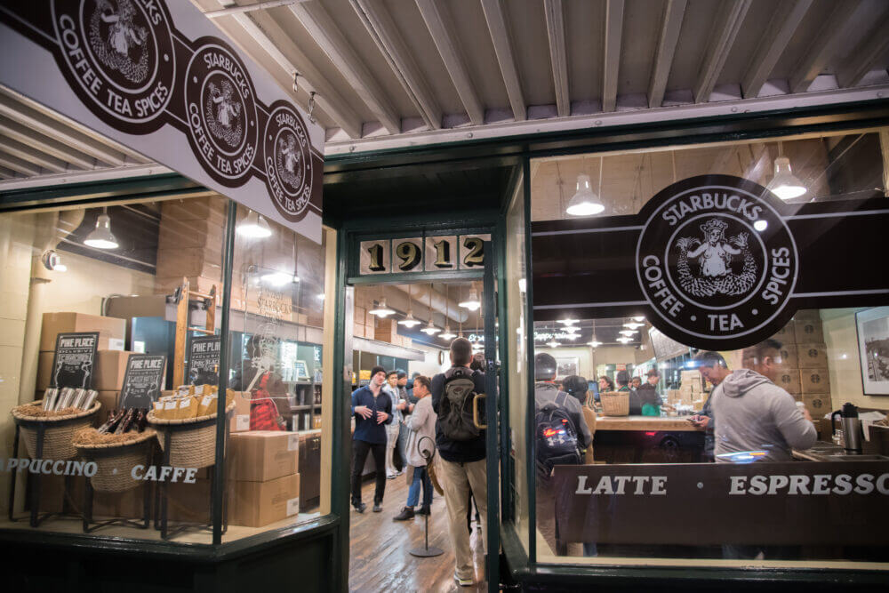 Close-up of the original Starbucks storefront at Pike Place Market in Seattle, showcasing the iconic brown logo and rustic shopfront. This historic coffee shop is a popular stop for visitors and a fun photo opportunity when exploring Pike Market with kids.