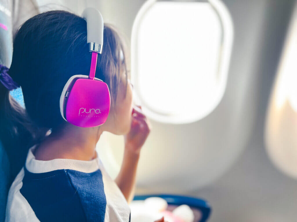 Flying with kids tips: Keep it simple and fun. Girl wearing PuroQuiet noise-canceling headphones while eating a snack, ready for a family vacation.