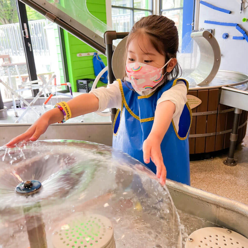 Toddler at KidsQuest Children’s Museum in Bellevue playing with the interactive water vapor bell exhibit, where water arcs up and over into a smooth bell shape—one of the museum’s engaging indoor experiences perfect for rainy or sunny days with kids.
