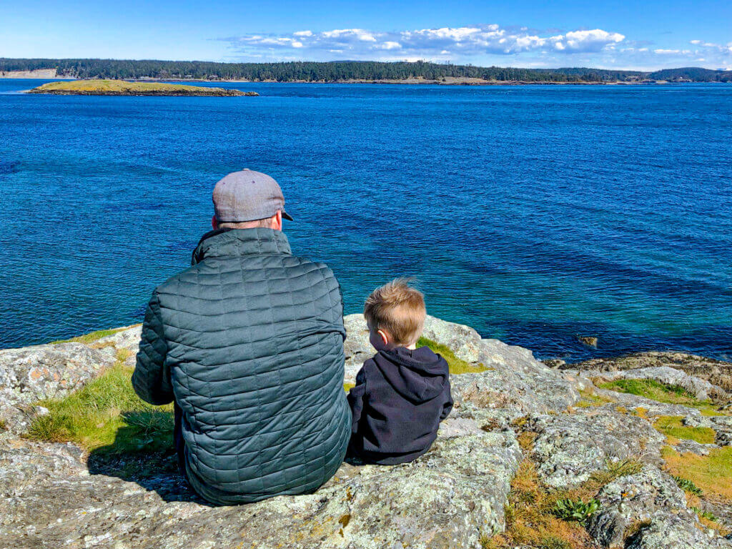 San Juan Island, Friday Harbor, with a dad and son sitting on rocks on a cliff looking out at the water views on a sunny summer day at Cattle Point.
