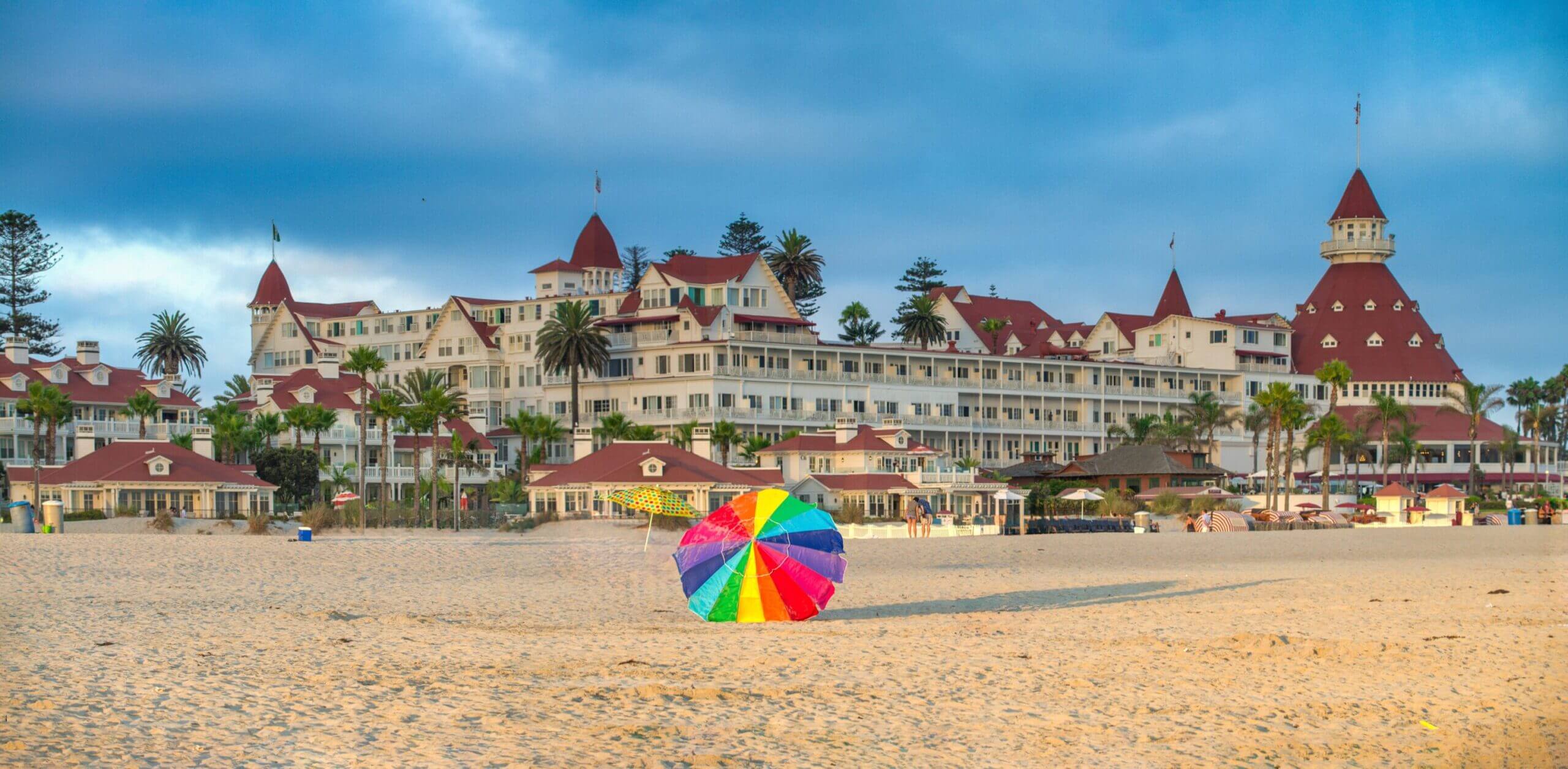 Panoramic view from the famous Coronado Beach, featuring a colorful rainbow umbrella on the sand with the historic Hotel del Coronado in the background. This kid-friendly beach is perfect for families, offering soft white sand, gentle waves, and plenty of space for children to run, build sandcastles, and splash in the water.