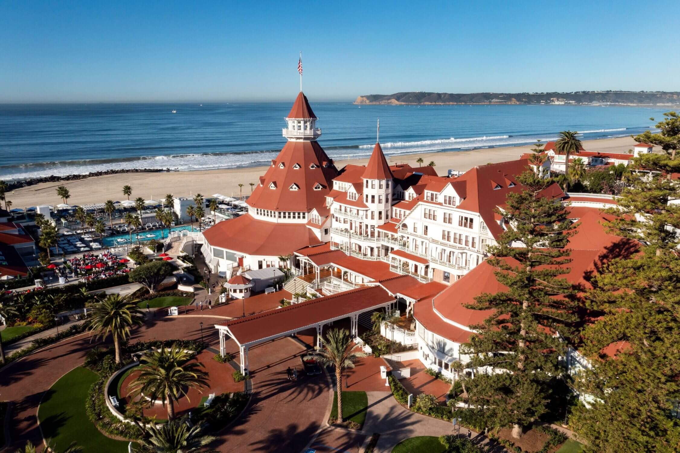 Aerial view of the iconic Hotel del Coronado, a historic beachfront resort on Coronado Island in San Diego. This stunning luxury hotel, known for its red-roofed Victorian architecture, sits along a pristine stretch of white sandy beach, offering breathtaking ocean views. A top destination for family vacations, Hotel del Coronado features world-class dining, beachfront activities, kid-friendly programs, and a rich history dating back to 1888.
