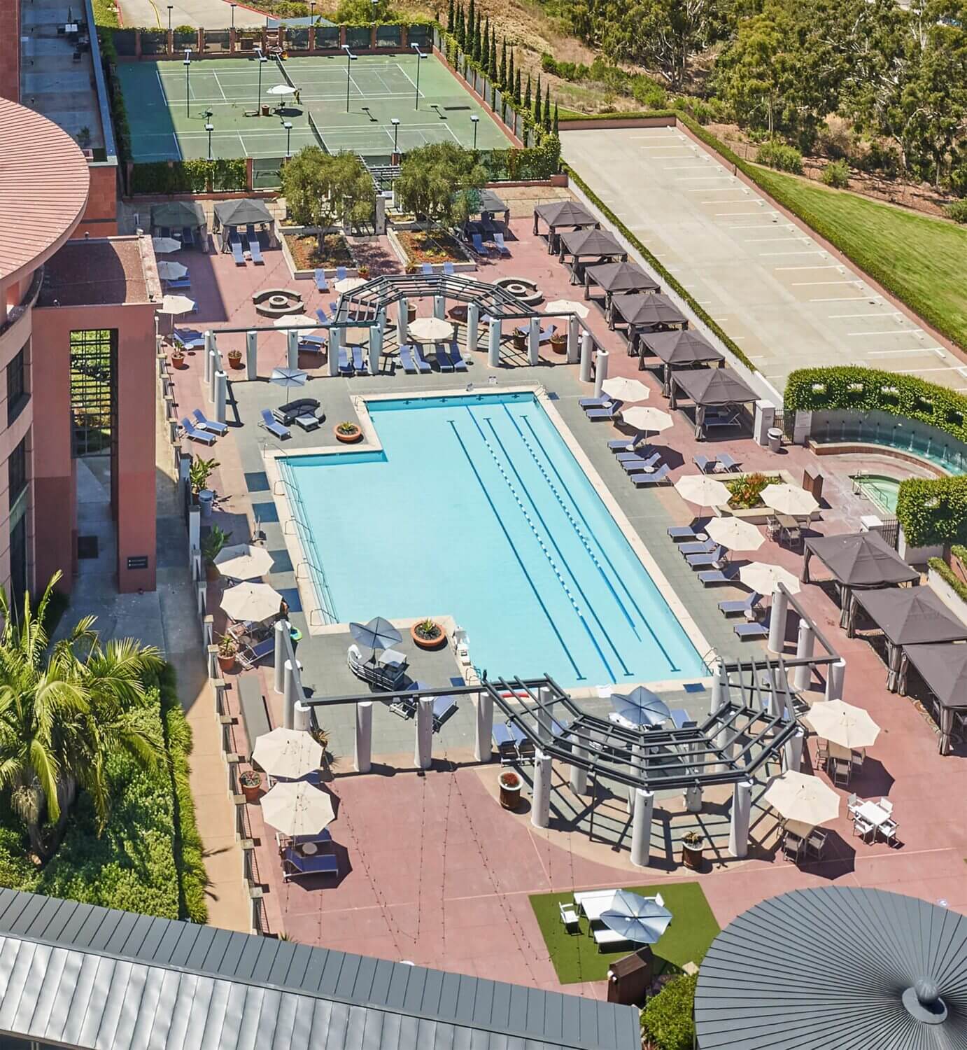 Aerial view of the outdoor swimming pool and tennis courts at Hyatt Regency La Jolla, showcasing a luxurious resort-style setting. The expansive heated pool is perfect for families, featuring plenty of lounge chairs, shaded cabanas, and a relaxing hot tub.