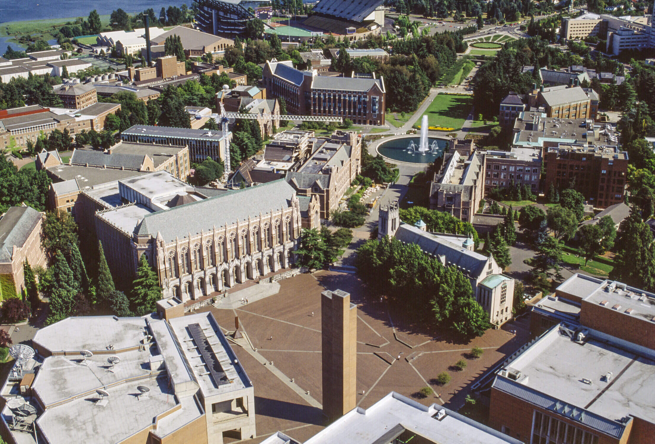 Aerial view of the University of Washington, featuring the iconic Suzzallo Library and the picturesque Drumheller Fountain. This stunning perspective captures the beauty of the UW campus, blending historic architecture, lush green spaces, and natural scenery in one panoramic image.