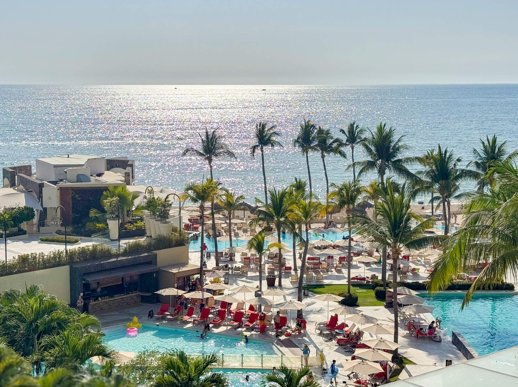 View from a hotel balcony at Dreams Vallarta Bay Resort, overlooking the sparkling pools, palm-lined grounds, and golden beachfront with ocean views stretching into the horizon. Some premium rooms in the Preferred Club section offer upgraded features like private hot tubs on the balcony and personalized butler service—perfect for relaxing while the kids play or winding down after a day of adventure. This stunning view showcases the resort’s tropical beauty and thoughtful amenities that make it a top choice for family-friendly luxury in Puerto Vallarta.