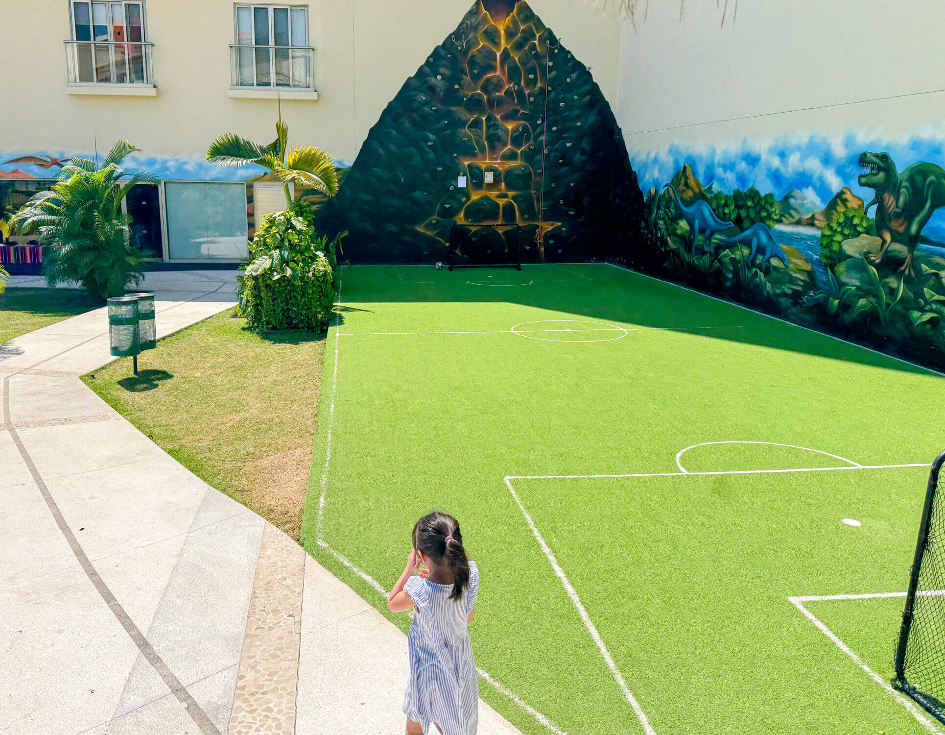 A young girl walks toward the indoor section of the Dreams Vallarta Bay Resort Kids Club, where children can color, play games, and enjoy air-conditioned fun. On her way, she passes the outdoor play area featuring a mini soccer field and kid-sized rock climbing wall—both popular activities among young guests. This supervised club offers a mix of creative play and outdoor adventure, giving kids a space to make friends while parents enjoy a little downtime at this family-friendly all-inclusive resort