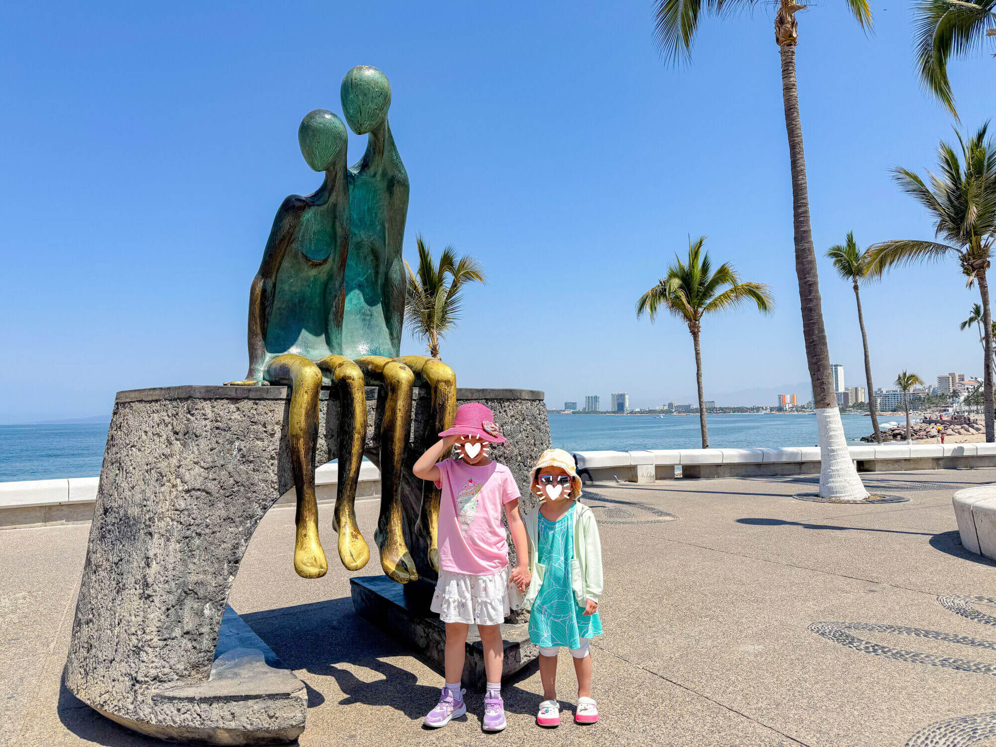 Two young girls pose happily in front of a whimsical art sculpture along the Puerto Vallarta Malecón, with stunning ocean views in the background. The boardwalk is lined with creative public art and makes for a fun, kid-friendly outing filled with photo ops, sea breezes, and cultural charm