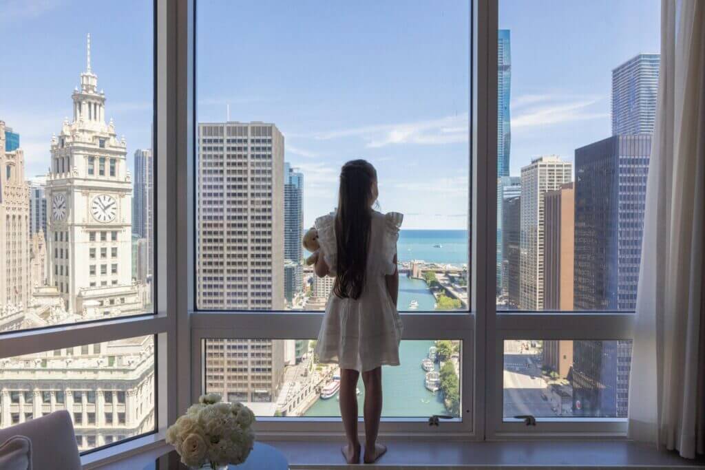 Young girl gazing out the window of a Chicago hotel room, with a scenic view of the Chicago River and iconic downtown architecture—highlighting one of the top kid-friendly hotels featured in our blog.