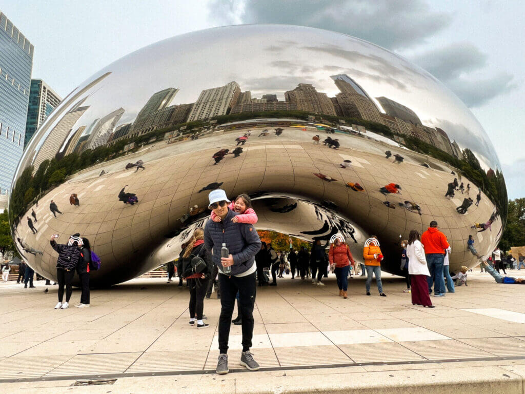 A dad and daughter pose for a photo in front of Chicago’s famous Cloud Gate sculpture, also known as “The Bean,” with city buildings reflected in the shiny surface—one of the must-see attractions in Chicago for families.