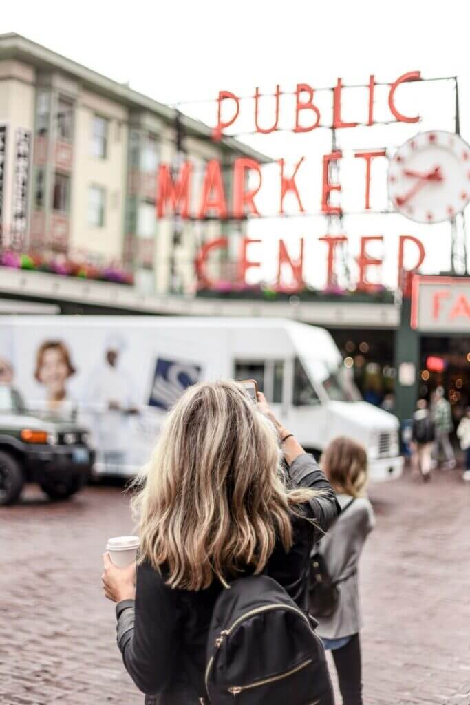 Seattle’s Pike Place Market – Mom tourist snapping a photo of the iconic market sign while holding a coffee, with a child slightly blurred in the foreground, capturing a classic family travel moment.