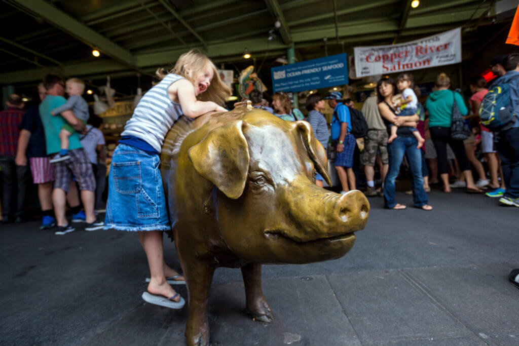 Rachel the Piggy Bank statue at Pike Place Market with a little girl happily hanging onto the bronze pig—this iconic photo spot is a must-do for families visiting Seattle, and a fun way for kids to interact with the market’s quirky charm.