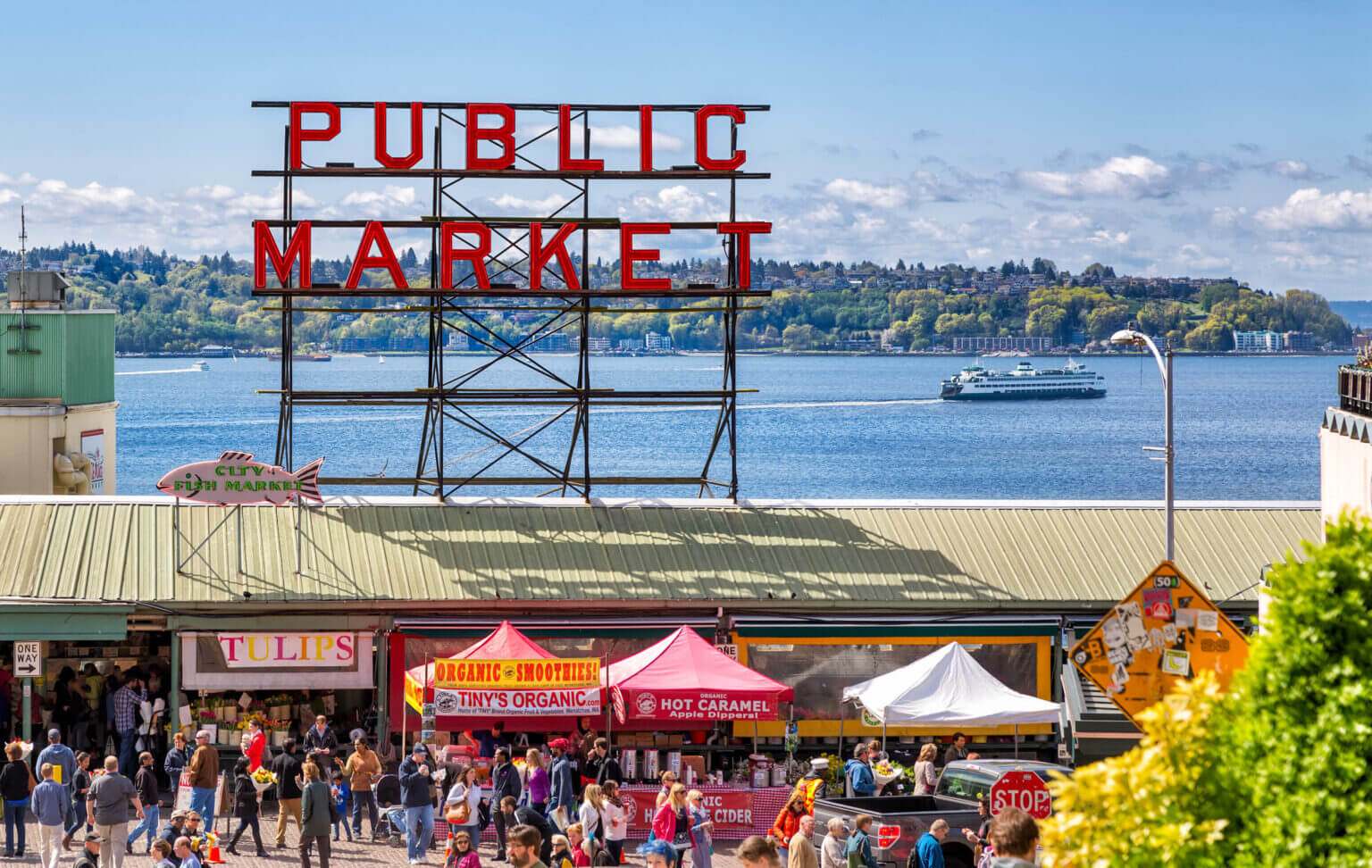 Pike Place Market on a sunny day with the iconic Public Market Center sign, bustling with tourists and families exploring the vendor stalls, with Puget Sound visible in the background — featured in our blog about top things to do with kids in Seattle.